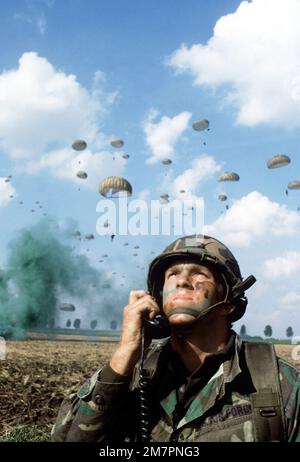 Ein Mitglied des Kampfkontrollteams in einer Abwurfzone leitet ein sich näherndes C-141-Starlifter-Flugzeug mit einem Funk in Position für einen Truppenabwurf. Im Hintergrund sind Truppen, die vom vorhergehenden Starlifter abspringen, am Boden. Der Flieger ist ein 317. taktisches Air-Wing-Mitglied, das an Übung Reforger '80 beteiligt ist. Betreff Betrieb/Serie: REFORGER '80 Land: Deutschland / Deutschland (DEU) Stockfoto