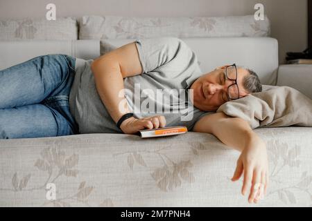 Ein alter Mann mit Brille schläft ein und liest ein Buch auf einer bequemen Couch im Wohnzimmer. Tagsüber schlafen, schlafen. Stockfoto