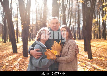 Portrait einer glücklichen dreiköpfigen Familie, die sich im sonnigen Wald umarmt. Eltern und Kinder im Teenageralter gehen im Herbstpark spazieren. Stockfoto