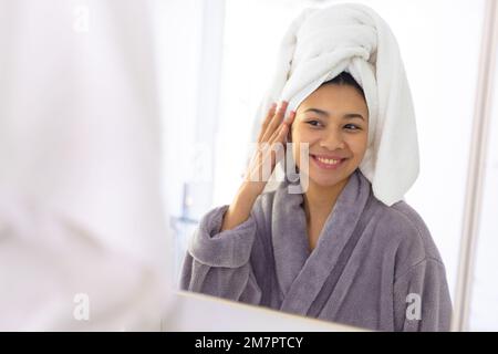 Glückliche, birassische Frau mit Bademantel und Handtuch, die im Badezimmerspiegel lächelt, mit Kopierbereich Stockfoto