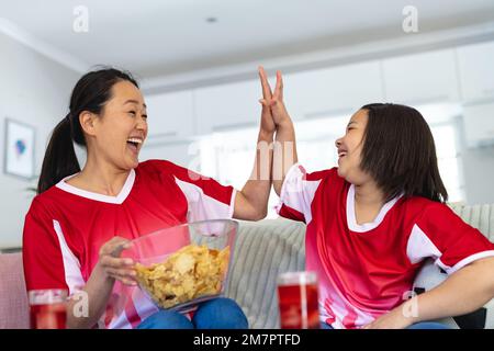 Glückliche asiatische Mutter und Tochter in Fußballtrikots, die sich Fußballspiele anschauen, Unterstützung leisten und High Fiving Stockfoto