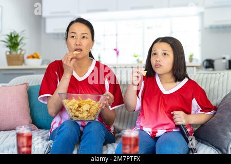 Asiatische Mutter und Tochter in Fußballtrikots, die Fußballspiele anschauen, Chips unterstützen und essen Stockfoto