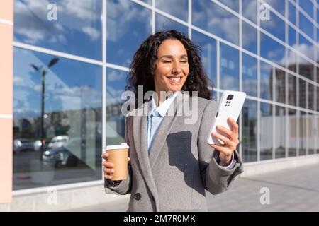 Wunderschöne und erfolgreiche hispanische Geschäftsfrau, die ihr Smartphone außerhalb des modernen Bürogebäudes benutzt, Frau mit einer Tasse heißes Getränk, die die Straße entlang läuft. Stockfoto