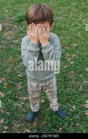 Ein kleiner Junge, der am St. Patrick's Day sein Gesicht mit Händen im Garten verdeckt Stockfoto