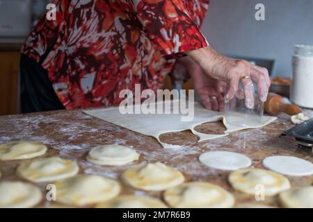 Nicht wiedererkennbare alte Frau schneidet Blätterteig mit einem Glas, um Knödel zuzubereiten. Spanien Stockfoto