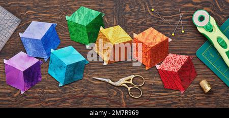 Diamanten, die in Form eines Würfels in den Farben des Regenbogens genäht sind, Quilt- und Nähzubehör auf Holzhintergrund. Stockfoto