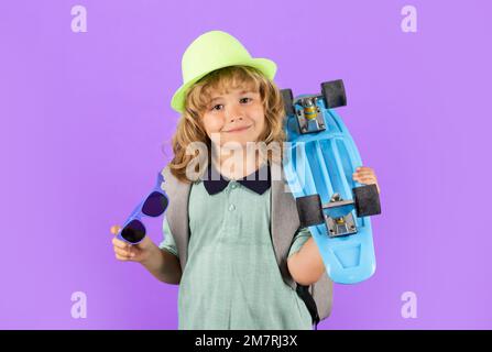 Ein Junge, der Longboard auf violettem isoliertem Hintergrund hält. Ein Junge mit Pennyboard. Studiofoto eines fröhlichen kleinen Kindes mit Penny-Board in modischer Mütze und Stockfoto