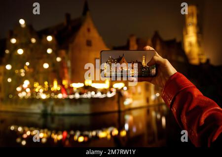 Fotografieren Sie die belgische Altstadt von Brügge, die bei Nacht beleuchtet ist. Tourist mit Smartphone Stockfoto