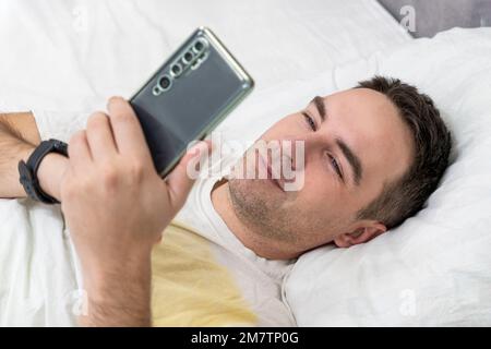 Ein fröhlicher Mann wachte morgens in seinem Schlafzimmer auf, benutzte ein Smartphone, Draufsicht. Ein Mann lächelt im Bett und sieht auf sein Smartphone. Stockfoto