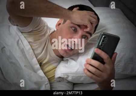 Gestresster junger Mann mit emotionalem Ausdruck und offenem Mund, der das Telefon hält und auf dem Smartphone auf die Weckzeit schaut, während er im Bett in Bedro liegt Stockfoto