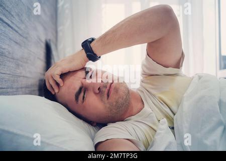 Der junge Mann hat Kopfschmerzen im Bett im Heim. Ein unverschlafener Kerl liegt im Bett mit einem unzufriedenen Gefühl im Gesicht. Morgens früh aufstehen Stockfoto