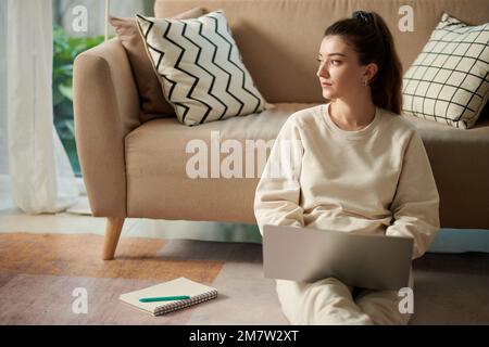 Seriöse junge Frau in Loungewear, die zu Hause auf dem Boden sitzt und an einem Laptop arbeitet Stockfoto