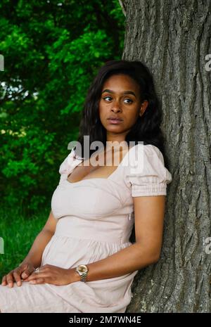 Eine junge Frau in einem weißen Kleid, die sich gegen eine Eiche lehnt. Stockfoto