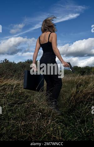 Eine junge blonde Frau, die schwarz trägt und eine Waffe (Pistole) und einen Aktenkoffer im Grasland in der Nähe von Wäldern hält. Thriller-Buch-Cover-Stil. Stockfoto