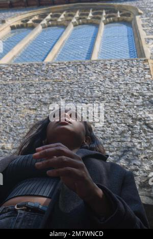 Ein Blick aus einem niedrigen Winkel auf eine junge Frau, die vor einer Kirchenmauer steht, mit einem Fenster darüber. Stockfoto