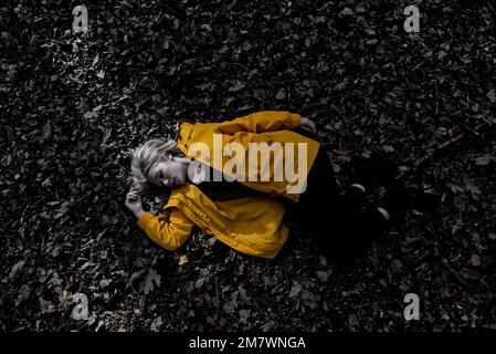 Eine junge blonde Frau, die einen gelben Mantel trägt, der auf dem Waldboden zwischen den Herbstblättern liegt. Thriller-Buch-Cover-Stil. Stockfoto