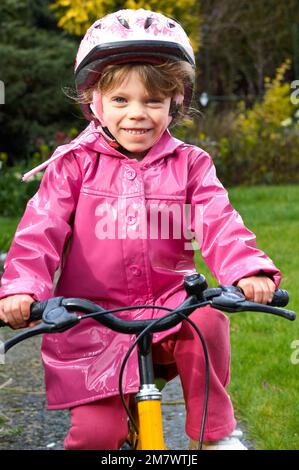 Ein glückliches 5-jähriges Mädchen, das in einem gelben Dreirad in einem pinkfarbenen, wasserdichten Mantel in einem Garten einen Weg hinunter fährt Stockfoto