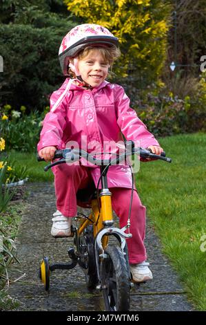 Ein glückliches 5-jähriges Mädchen, das in einem gelben Dreirad in einem pinkfarbenen, wasserdichten Mantel in einem Garten einen Weg hinunter fährt Stockfoto