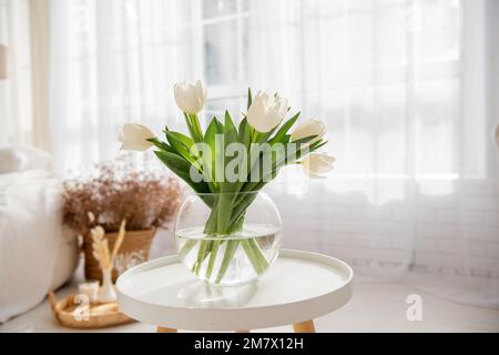 Auf einem runden Holztisch stehen Glasvase mit einem Strauß weißer Tulpen. Luftige, helle Einrichtung in hellen Farben. Frische, Reinheit natürlicher Farben. Sp Stockfoto