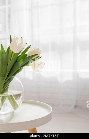 Auf einem runden Holztisch stehen Glasvase mit einem Strauß weißer Tulpen. Luftige, helle Einrichtung in hellen Farben. Frische, Reinheit natürlicher Farben. Sp Stockfoto