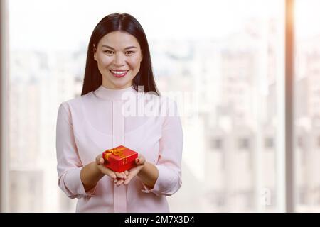 Lächelnde asiatische Frau, die eine rote Geschenkbox schenkt. Hintergrund für verschwommene Fenster. Stockfoto