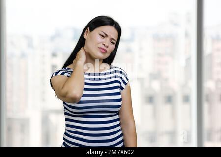 Porträt einer an Nackenschmerzen leidenden asiatischen Frau. Hintergrund von Office-Fenstern. Stockfoto