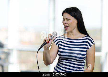 Junge brünette Asiatin singt ein Lied mit Mikrofon. Verschwommener heller Hintergrund. Stockfoto