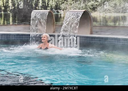 Fröhliche lächelnde, reife Frau mit kurzen grauen Haaren im Thermalbad im Freien mit Hydromassage. Konzept für aktive Senioren. Stockfoto