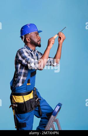 Ein afroamerikanischer Bauarbeiter auf der Leiter repariert etwas mit einem Schraubenzieher. Bauarbeiter trägt Uniform und Schutzhelm vor blauem Hintergrund bei Studioaufnahmen Stockfoto