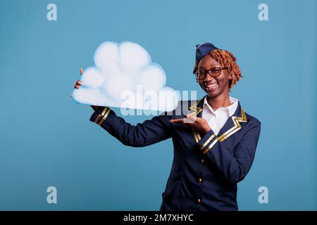 Weibliche Flugbegleiterin lächelt, während sie die Papierwolke vor blauem Hintergrund in der Studioaufnahme hielt. Optimistische Stewardess in Uniform, Konzept des Fliegens zwischen den Wolken. Stockfoto