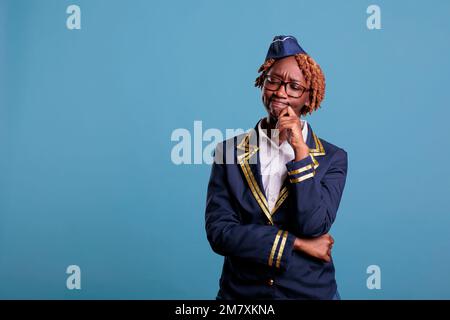 afroamerikanische Flugbegleiterin mit zweifelhaftem Gesichtsausdruck trägt Uniform vor blauem Hintergrund im Studio. Flugbegleiter denkt über die beste Flugdienststrategie nach. Stockfoto