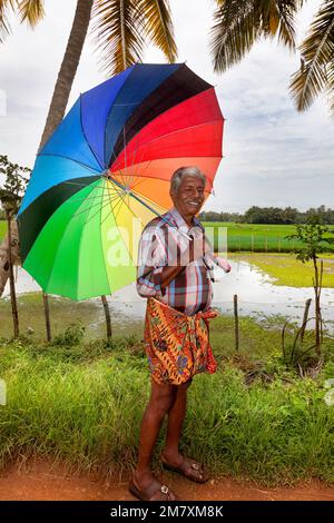 Allepey, Indien - 7. September 2012: Ein indischer Mann, gekleidet im typischen Kerala longui, ist mit einem großen bunten Daumen vor den Monsunregen geschützt Stockfoto