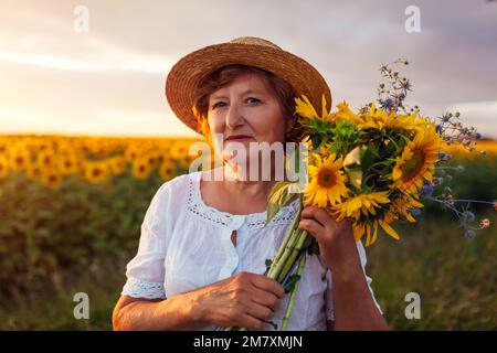 Eine ältere Frau hält bei Sonnenuntergang einen Strauß gelber Sonnenblumen auf dem Sommerfeld. Portrait Og, eine Frau mittleren Alters mit Hut Stockfoto