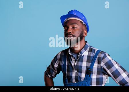 Müde erschöpfte Bauarbeiter in Bauhelm und Overalls. afroamerikanischer Angestellter mit geschlossenen Augen nach harter Arbeit, isoliert auf blauem Hintergrund, ermüdet. Stockfoto