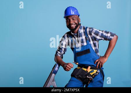 Der Baumeister sieht bei der Arbeit glücklich und entspannt aus, trägt Overalls und einen Schutzhelm, der sich auf eine tragbare Leiter lehnt. Optimistischer Bauarbeiter trägt Werkzeuggürtel während einer Pause. Stockfoto