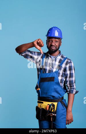 Studioporträt eines verärgerten Bauarbeiters mit Anzeichen von Missbilligung bei der Arbeit. Unzufriedener Mitarbeiter mit Schutzhelm und Arbeitskleidung vor einem isolierten blauen Hintergrund. Stockfoto