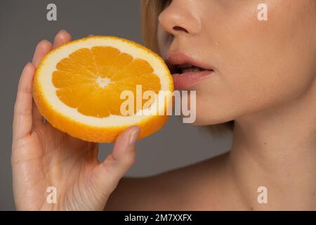 Zugeschnittene Ansicht einer jungen Frau, die eine saftige orangefarbene Hälfte in der Nähe weicher Lippen hält, isoliert auf einem grauen Stockbild Stockfoto