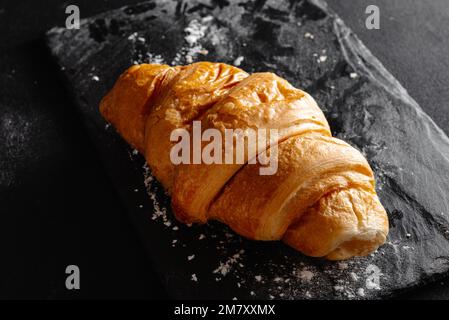 Großes Croissant auf schwarzem Hintergrund. Frisches und köstliches französisches Gebäck, Bäckereikonzept, Nahaufnahme Stockfoto