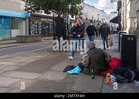 Ein obdachloser Mann, männlich; sitzt auf dem Bürgersteig, während Fußgänger im Stadtzentrum von Truro in Cornwall, England, im Vereinigten Königreich vorbeilaufen. Stockfoto