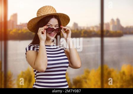 Porträt einer jungen Asiatin mit Strohhut und Sonnenbrille. Fenster mit Herbstlandschaft. Stockfoto