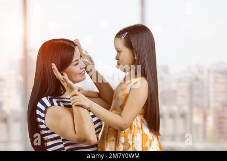 Die junge Mutter und ihre Tochter machen sich Gesichter. Das kleine Mädchen zeigt ihrer mutter Zunge. Hintergrund für verschwommene Fenster. Stockfoto