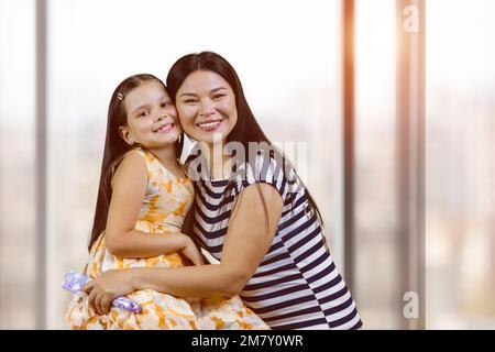 Porträt einer glücklichen asiatischen Frau mit ihrer adoptierten weißen Tochter. Süßes kleines Mädchen mit ihrer Mutter drinnen. Hintergrund für verschwommene Fenster. Stockfoto