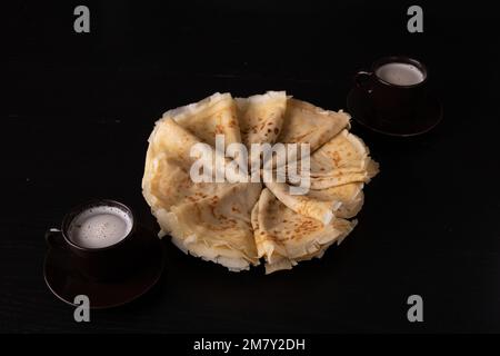 Fotografieren Sie Pfannkuchen auf einem Teller in einem Kreis mit zwei Tassen auf schwarzem Hintergrund Stockfoto
