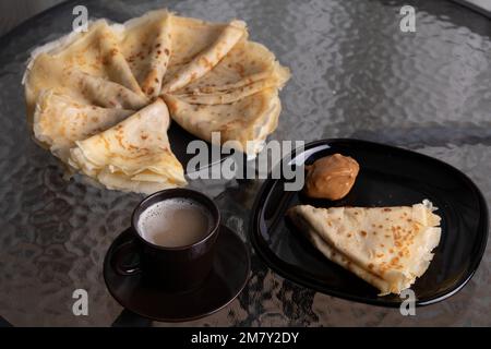 Fotografieren Sie jede Menge gebratene Pfannkuchen und abgekochte Kondensmilch auf einem Teller und eine Tasse Kaffee auf einem Glastisch Stockfoto