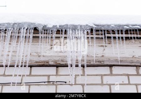 Schneebedecktes Dach eines Hauses mit Eiszapfen, Nahaufnahme auf dem Hintergrund einer weißen Ziegelwand Stockfoto