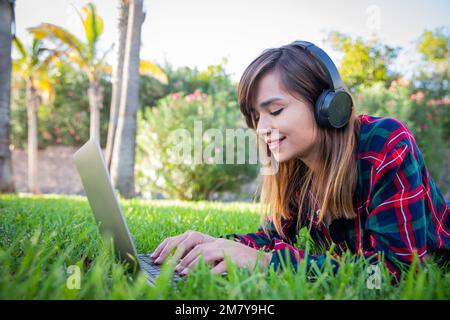 Eine junge Frau arbeitet mit ihrem Laptop und hört Musik, während sie im Garten liegt Stockfoto