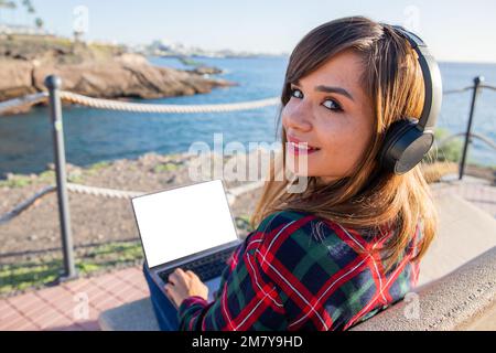 Ein Mädchen benutzt ihren Laptop am Meer, sitzt auf einer Bank und zeigt ein Modell Stockfoto