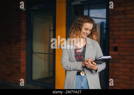 Außenporträt einer fröhlichen Studentin mit Arbeitsbüchern, die neben dem College-Gebäude stehen, nach unten schauen und lächeln. Lebensstil der Menschen. Ein glücklicher Lebensstil Stockfoto