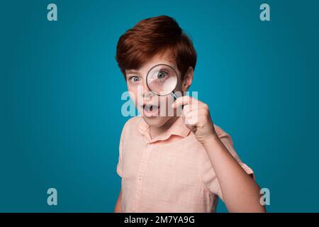 Ein verblüffter Junge, der durch eine Lupe blickt, isoliert auf blauem Hintergrund. Porträt eines süßen kleinen Jungen. Großes Auge. Stockfoto