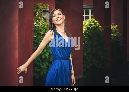 Multiethnische glückliche junge, lächelnde Frau in blauem Kleid, die auf der Sommerterrasse mit Säulen eines Luxushauses steht Stockfoto